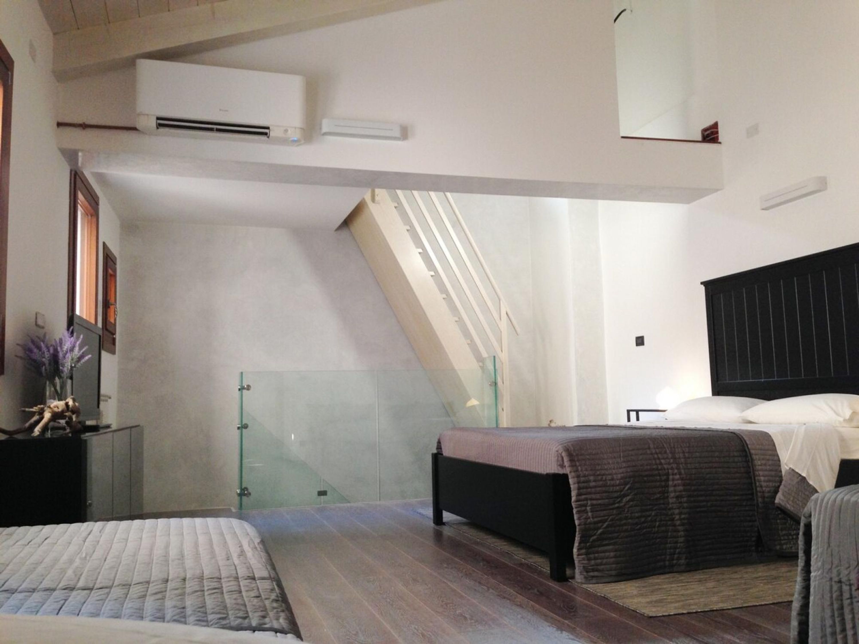 A bedroom with a sloped ceiling, a glass staircase, a bed, a bedside table, and an air conditioning unit.
