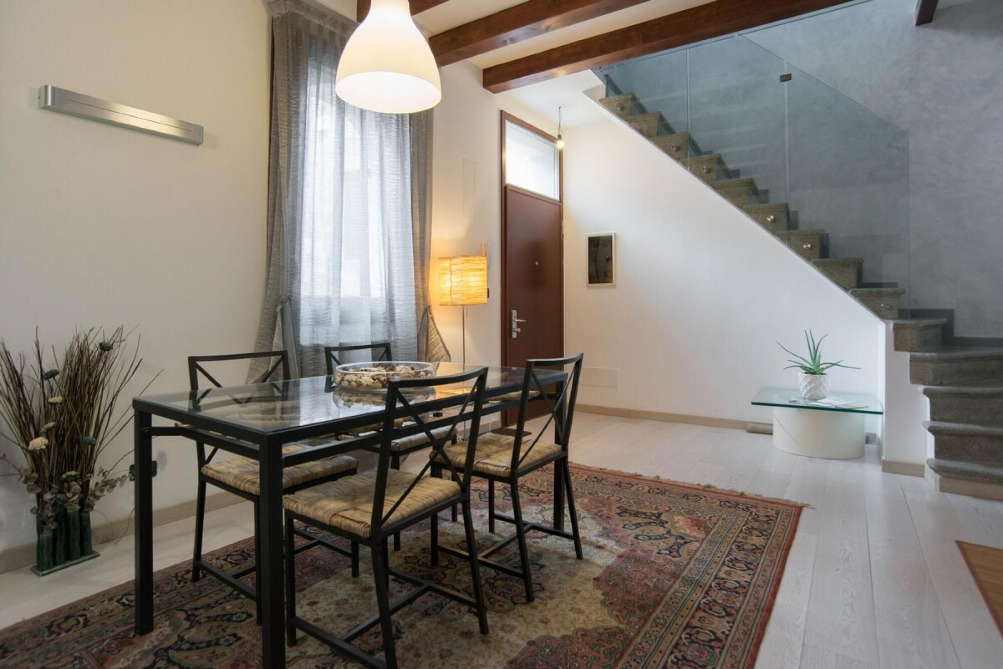 A dining area with a glass table and chairs, a patterned rug, and a staircase with a glass railing.