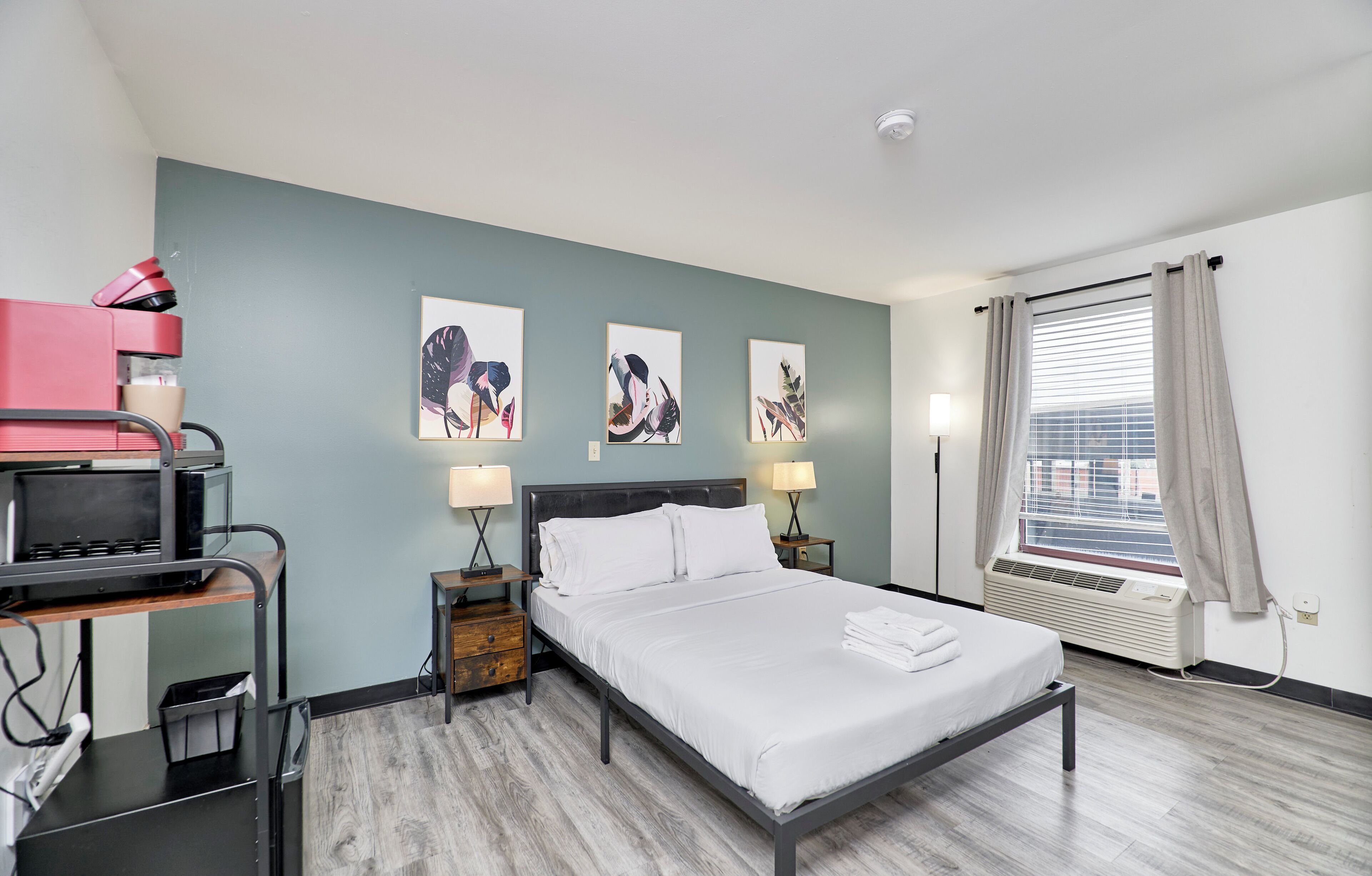 A hotel room with a bed, bedside lamps, a desk with a computer, and artwork on the wall.