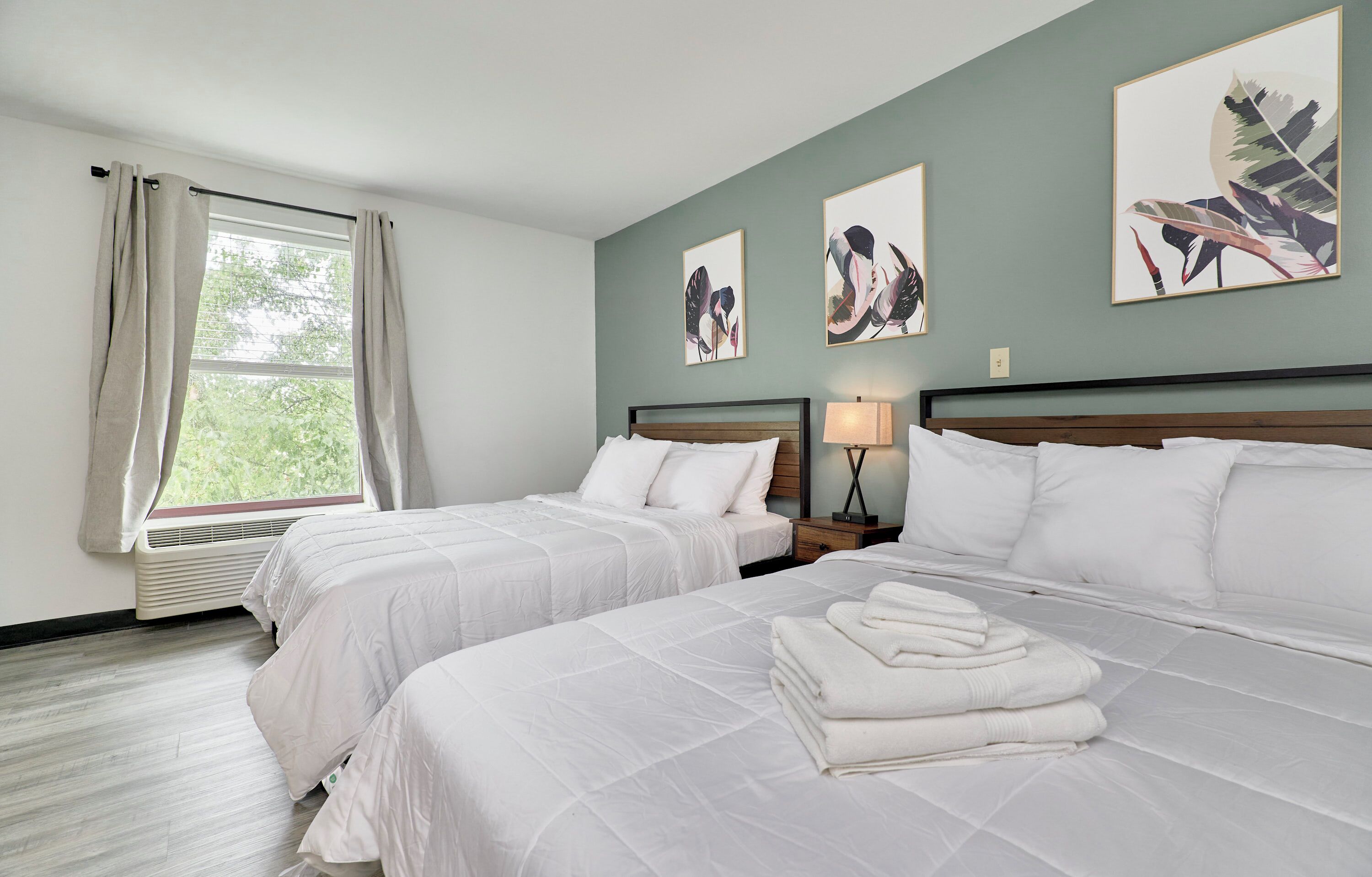 A hotel room with two beds, a window with curtains, a heater, and artwork on the walls.