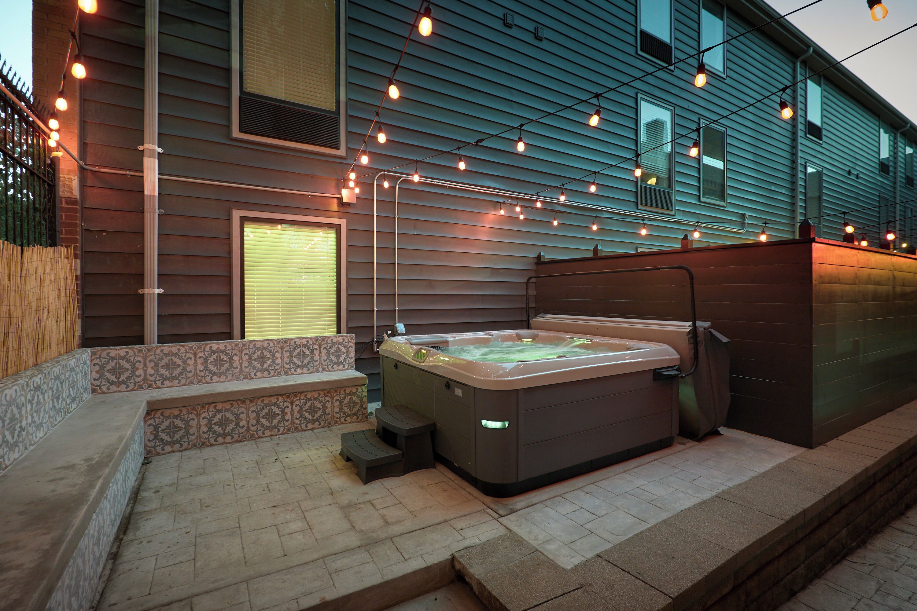 A hot tub in a backyard with a tiled patio, string lights, and a house with horizontal siding.