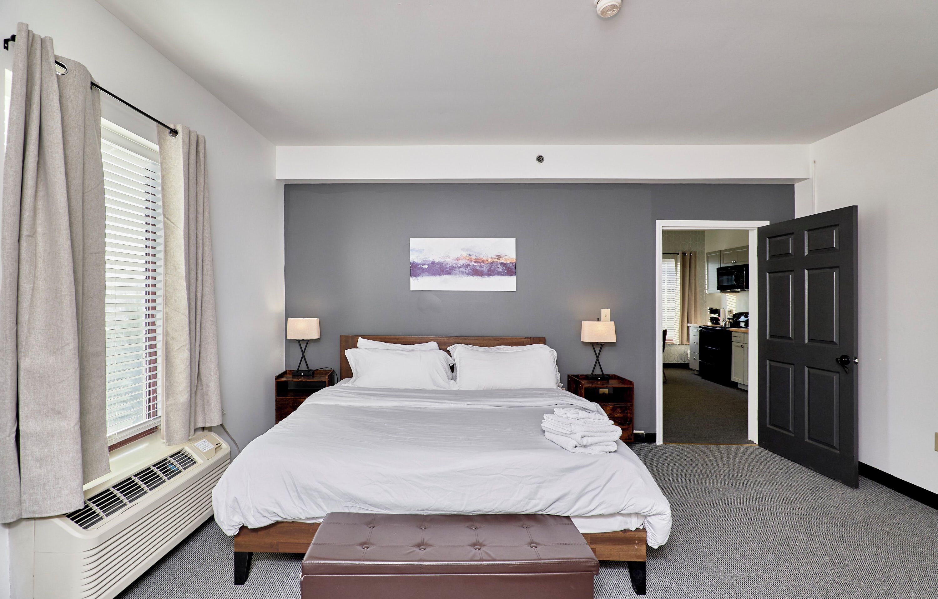 A hotel room with a large bed, two bedside tables with lamps, a window with curtains, a heating unit, and a door leading to a hallway.