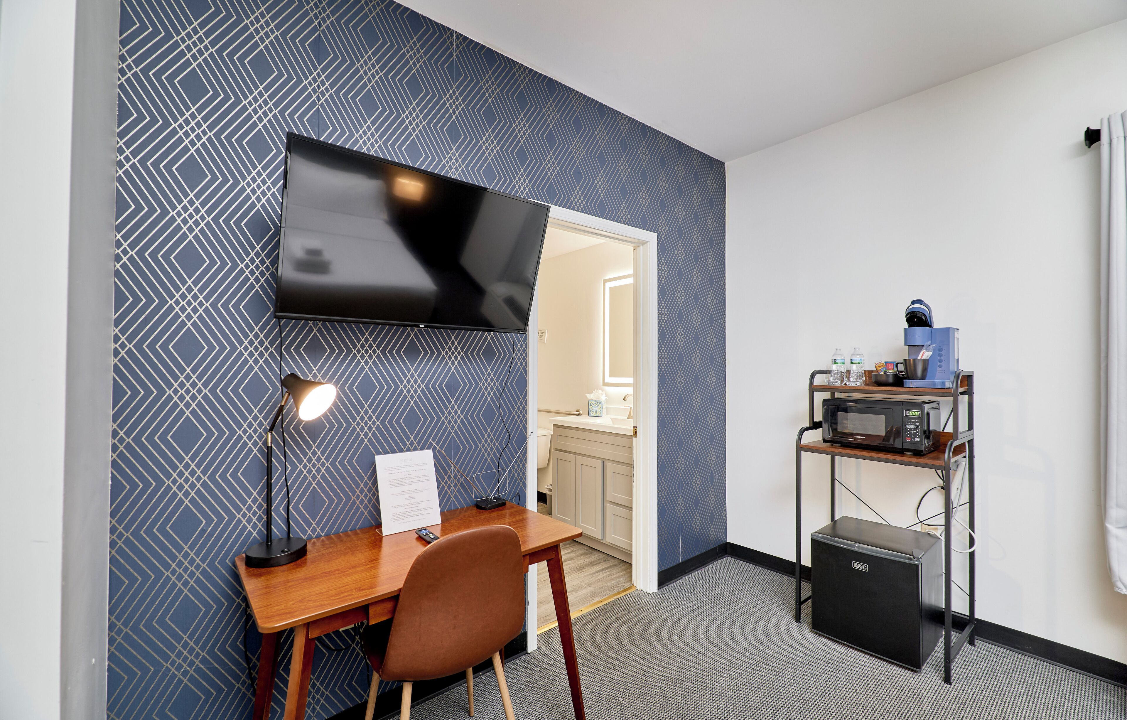 Une chambre d’hôtel équipée d’un bureau, d’une chaise, d’un téléviseur à écran plat, d’une lampe et d’un coin cuisine comprenant un four à micro-ondes et une machine à café.