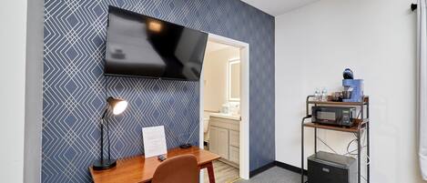 Une chambre d’hôtel équipée d’un bureau, d’une chaise, d’un téléviseur à écran plat, d’une lampe et d’un coin cuisine comprenant un four à micro-ondes et une machine à café.