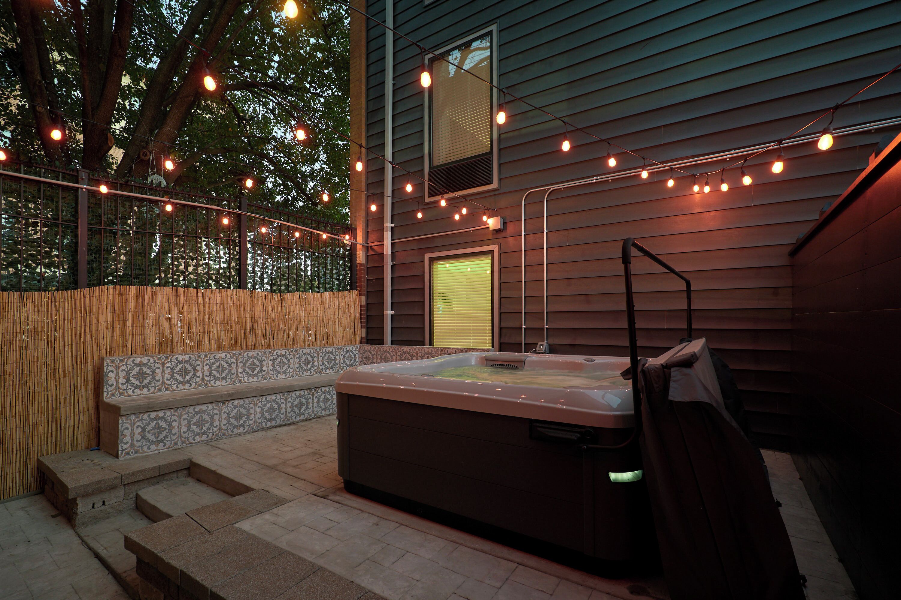Zona de hidromasaje exterior con escalones que dan acceso a ella, rodeada por una valla de madera y luces colgantes.