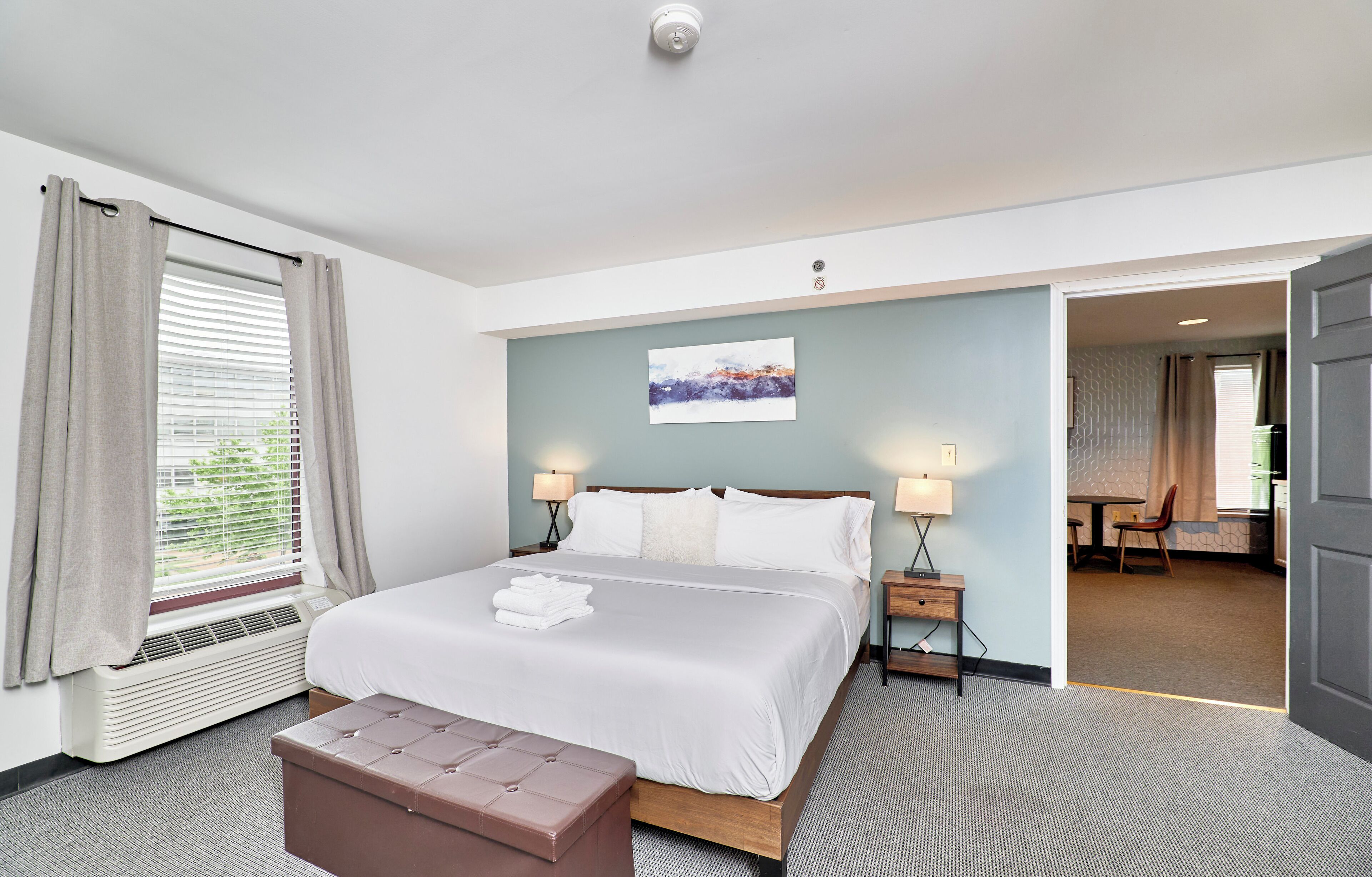 Une chambre d’hôtel avec un lit, des tables de chevet, un banc, une fenêtre avec des rideaux, un tableau au mur et une porte ouverte donnant sur une autre pièce.
