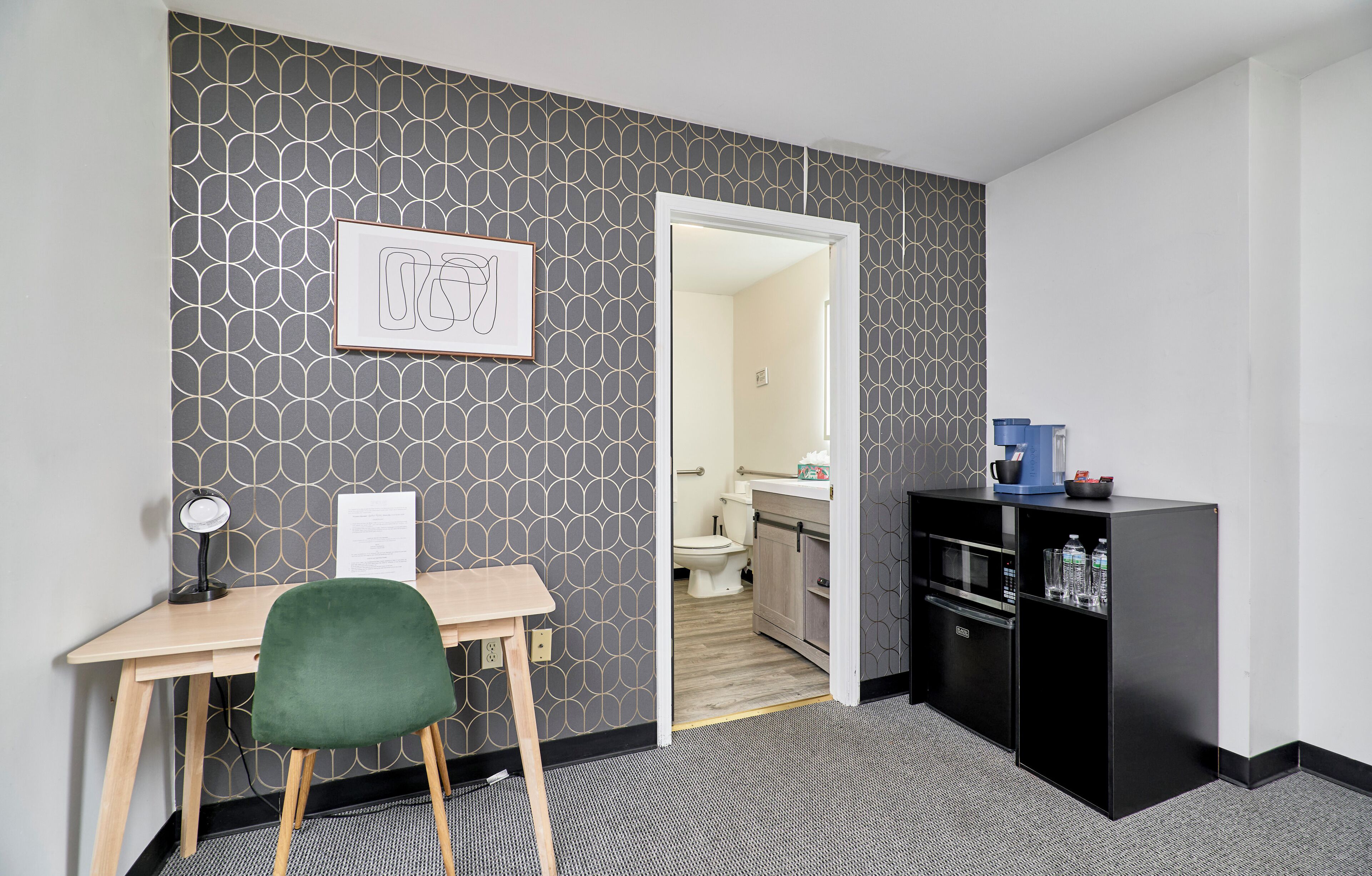 A hotel room with a desk, a green chair, a black minibar, and a bathroom visible through an open door.