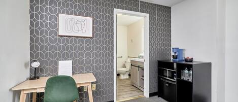 A hotel room with a desk, a green chair, a black minibar, and a bathroom visible through an open door.