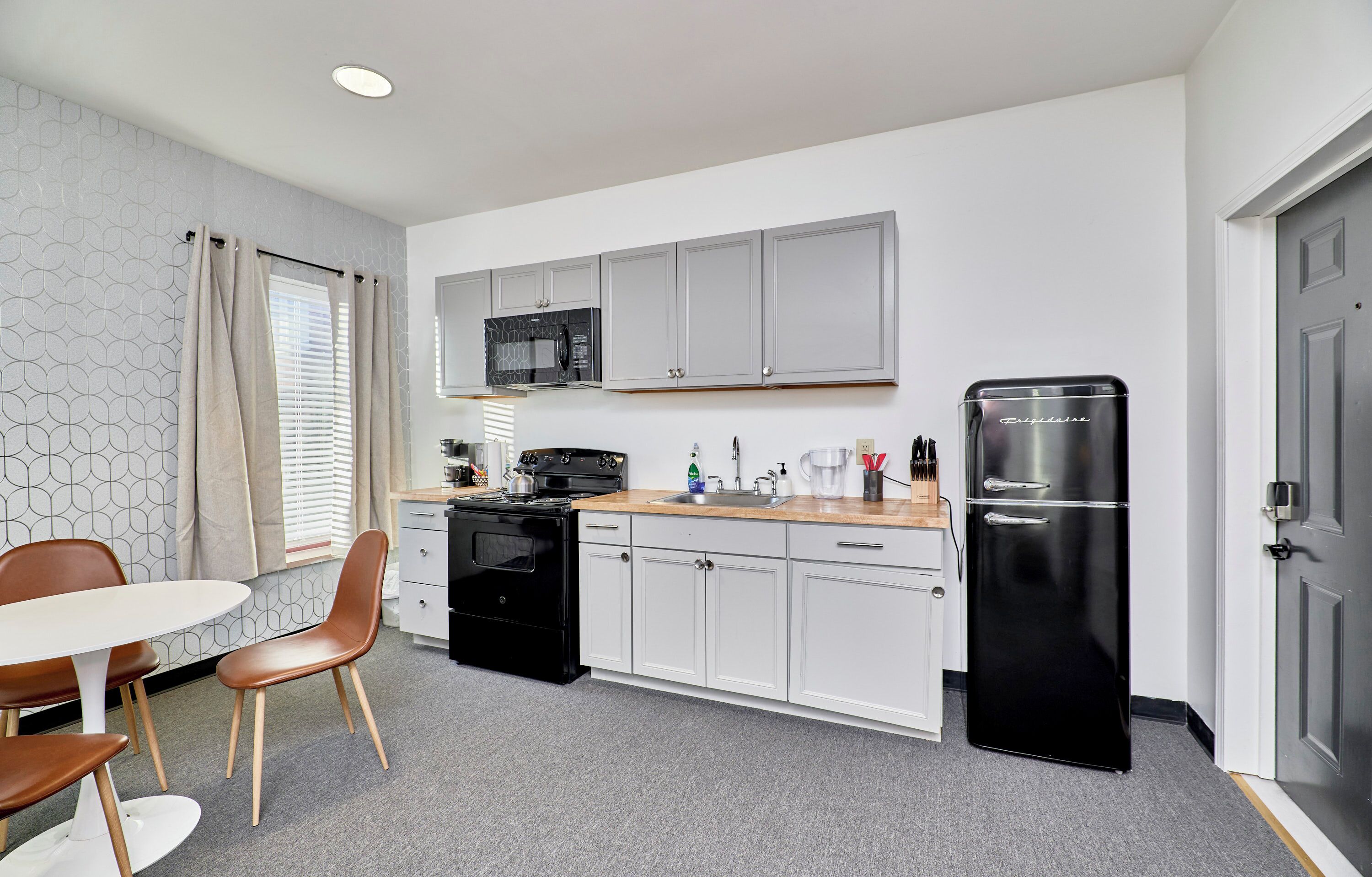 Cocina compacta con armarios blancos, un refrigerador negro, un microondas y una estufa. Hay una mesa de comedor con sillas y una ventana con cortinas.