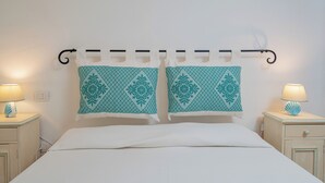 Una cama con ropa blanca, dos almohadas estampadas en color verde azulado y una mesita de noche con una lámpara.