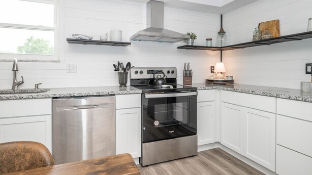 A modern kitchen with white cabinetry, a stainless steel dishwasher, a black oven, and a gas stove. There are shelves above the counter holding kitchen items and a window providing natural light. - StayBurgundy Fairfield, OH (Fairfield)