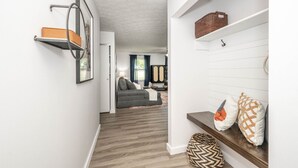A hallway with a wooden floor, a gray sofa in the background, and a white wall with a shelf holding a basket and a picture frame. - StayBurgundy Fairfield, OH (Fairfield)