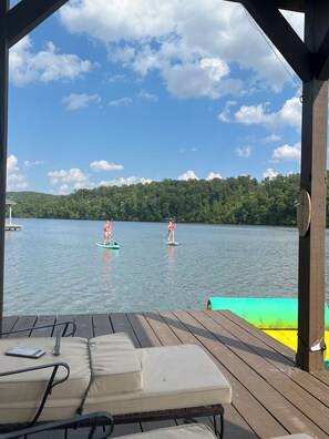 Una terraza de madera con sillones de descanso y vistas a dos personas practicando paddleboard en un lago.