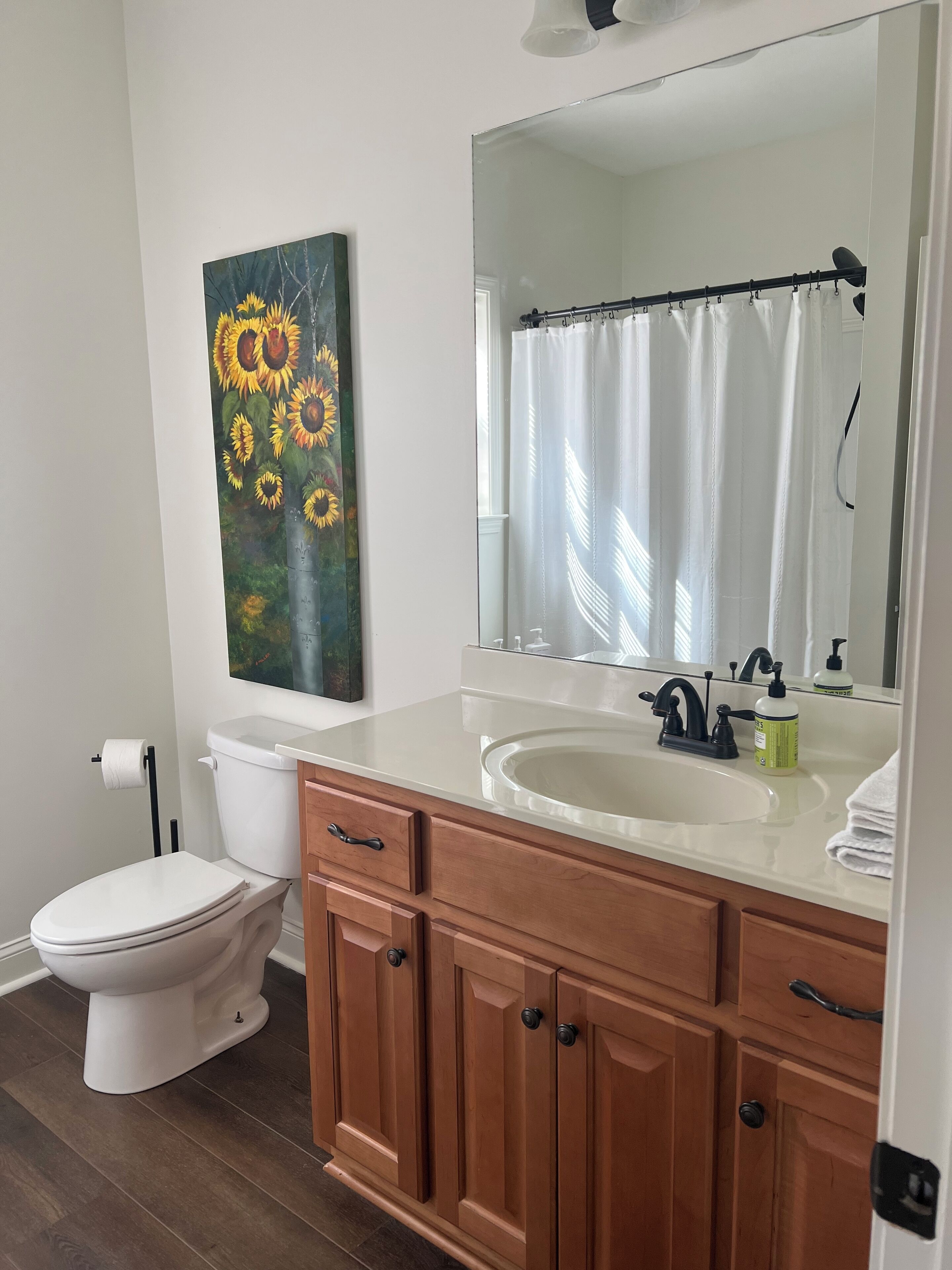 Une salle de bain avec des toilettes blanches, un meuble-lavabo en bois avec un évier, un grand miroir et un tableau représentant des tournesols accroché au mur.