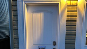 A white door with an 'Airbnb' sign, a small porch with a railing, and a green hose on the ground. - Brand new 2 bedroom basement in Leduc(10mins from YEG airport) (Leduc)