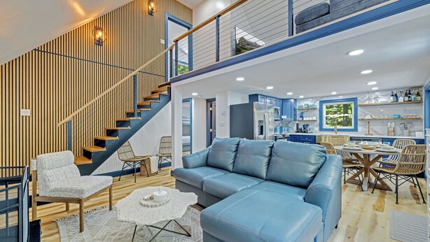 A modern living room with a blue sectional sofa, a wooden staircase, and a kitchen area with blue cabinets and a dining table. - Charming /Lake/Kayak/Firepit/Hot tub/Pets (Blakeslee)