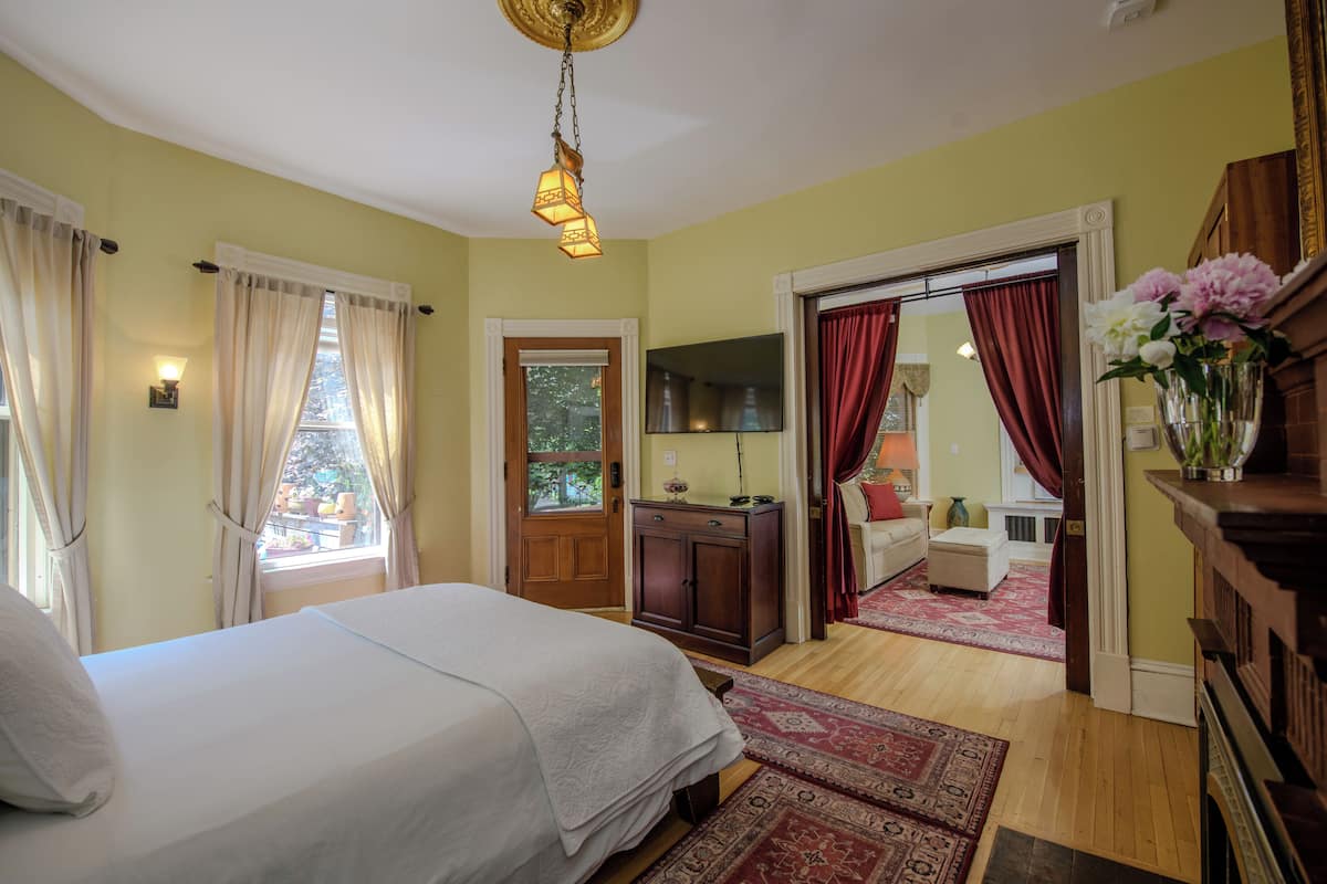 Een slaapkamer met een bed, een houten dressoir, een bureau en een stoel. Er is een raam met gordijnen, een deur en een spiegel.