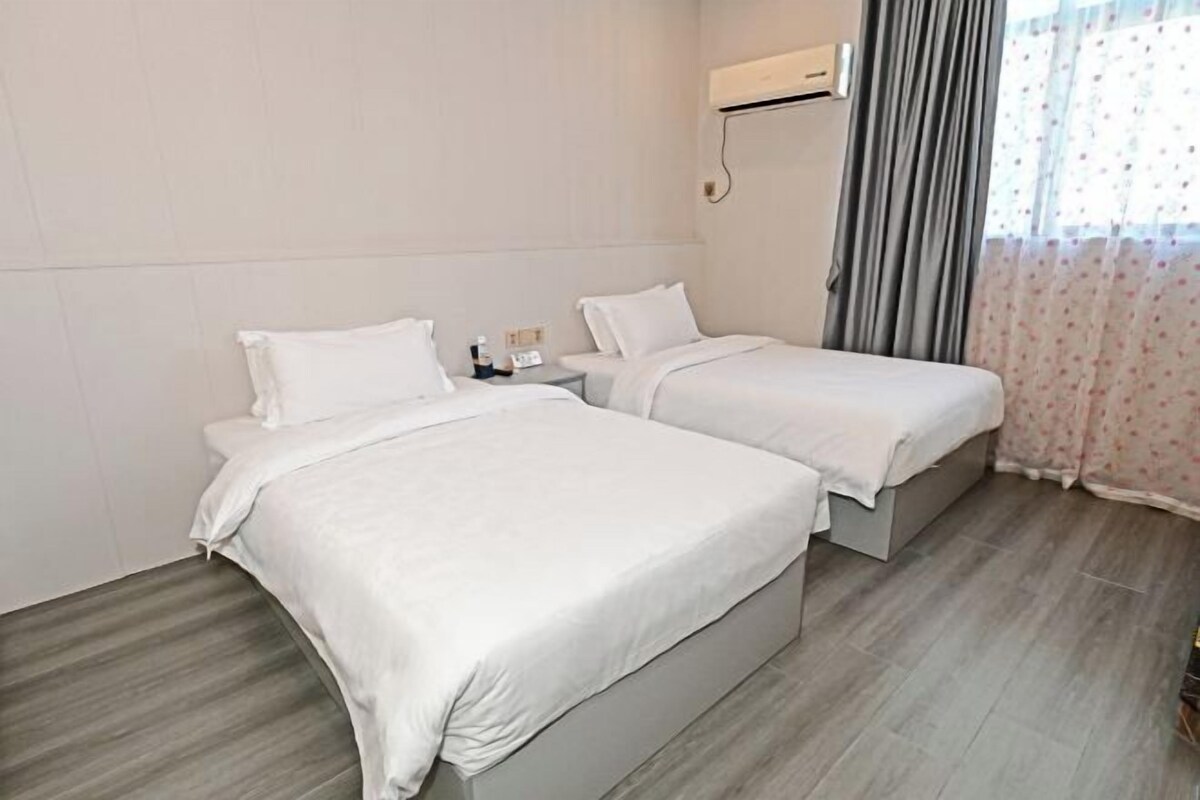A hotel room with two beds, a nightstand, a wall-mounted air conditioner, and a window with curtains.