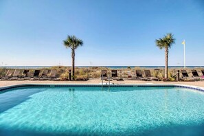 A clear blue pool with lounge chairs and palm trees, overlooking a beach.