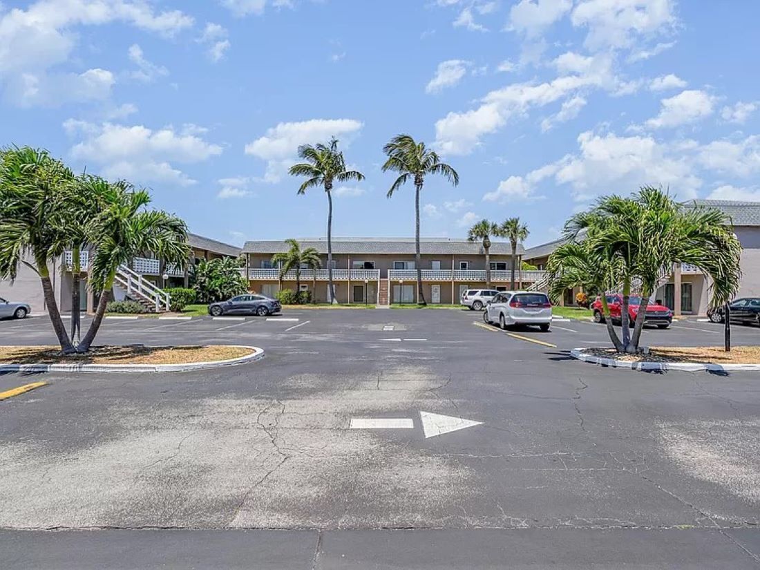 A parking lot with cars, palm trees, and a building with a flat roof in the background.