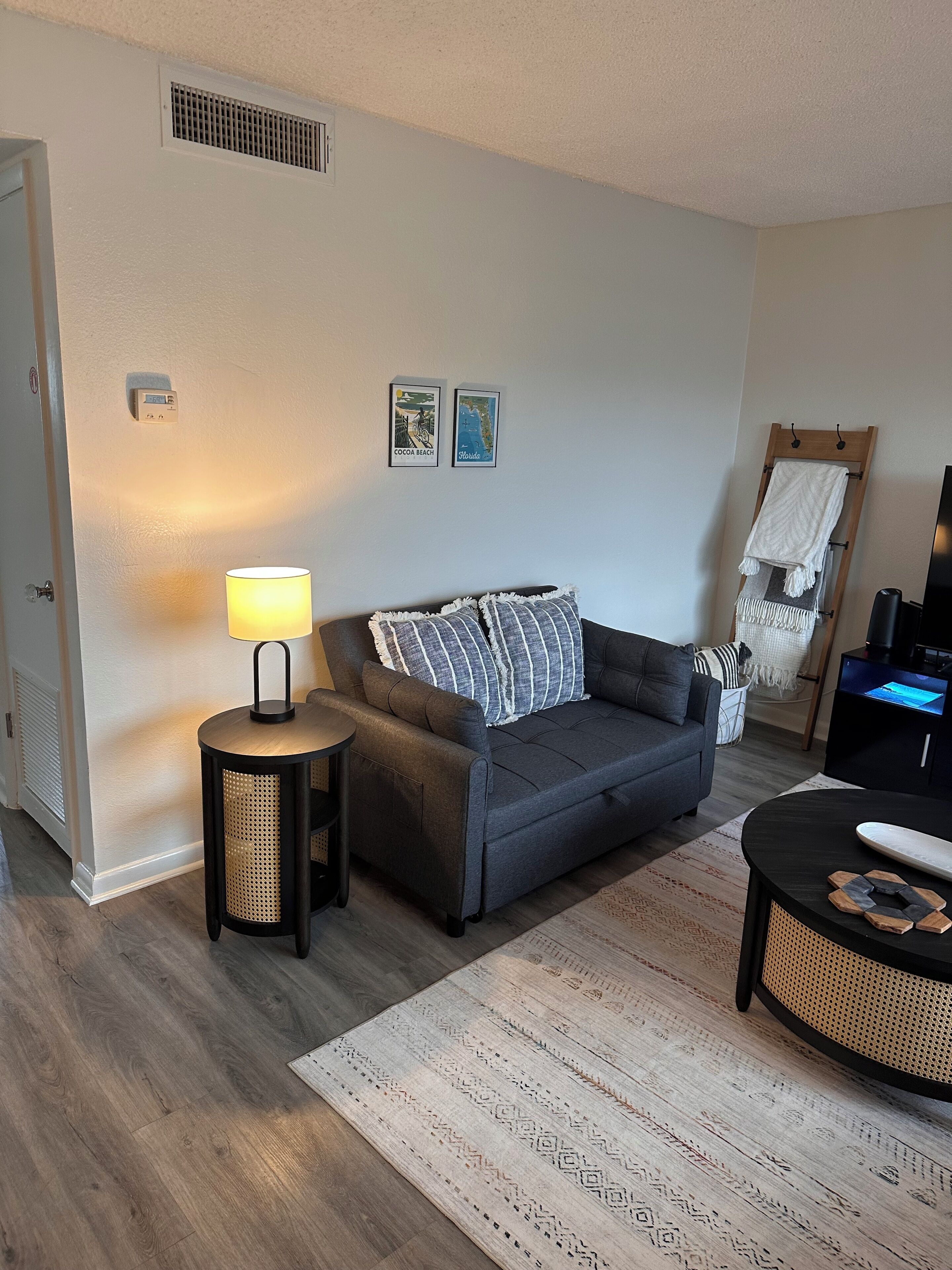 Una sala de estar con un sofá gris, una mesita redonda con lámpara, una alfombra estampada y un televisor sobre un mueble bajo.