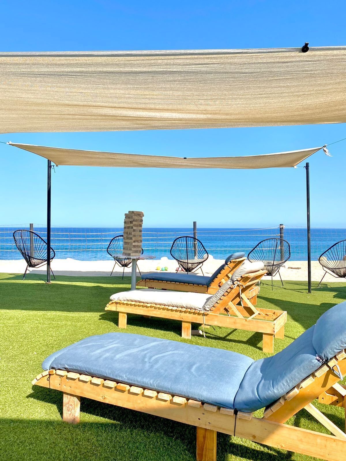 A beachside lounge area with wooden loungers and wire mesh chairs, a wooden table, and a view of the ocean.