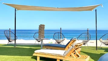 Eine Strandlounge mit Holzliegen und Stühlen aus Drahtgeflecht, einem Holztisch und Blick auf den Ozean.