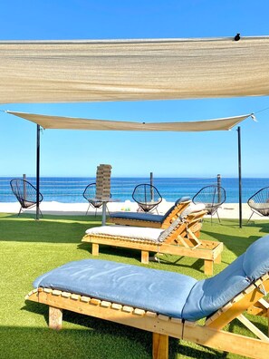 Eine Strandlounge mit Holzliegen und Stühlen aus Drahtgeflecht, einem Holztisch und Blick auf den Ozean.