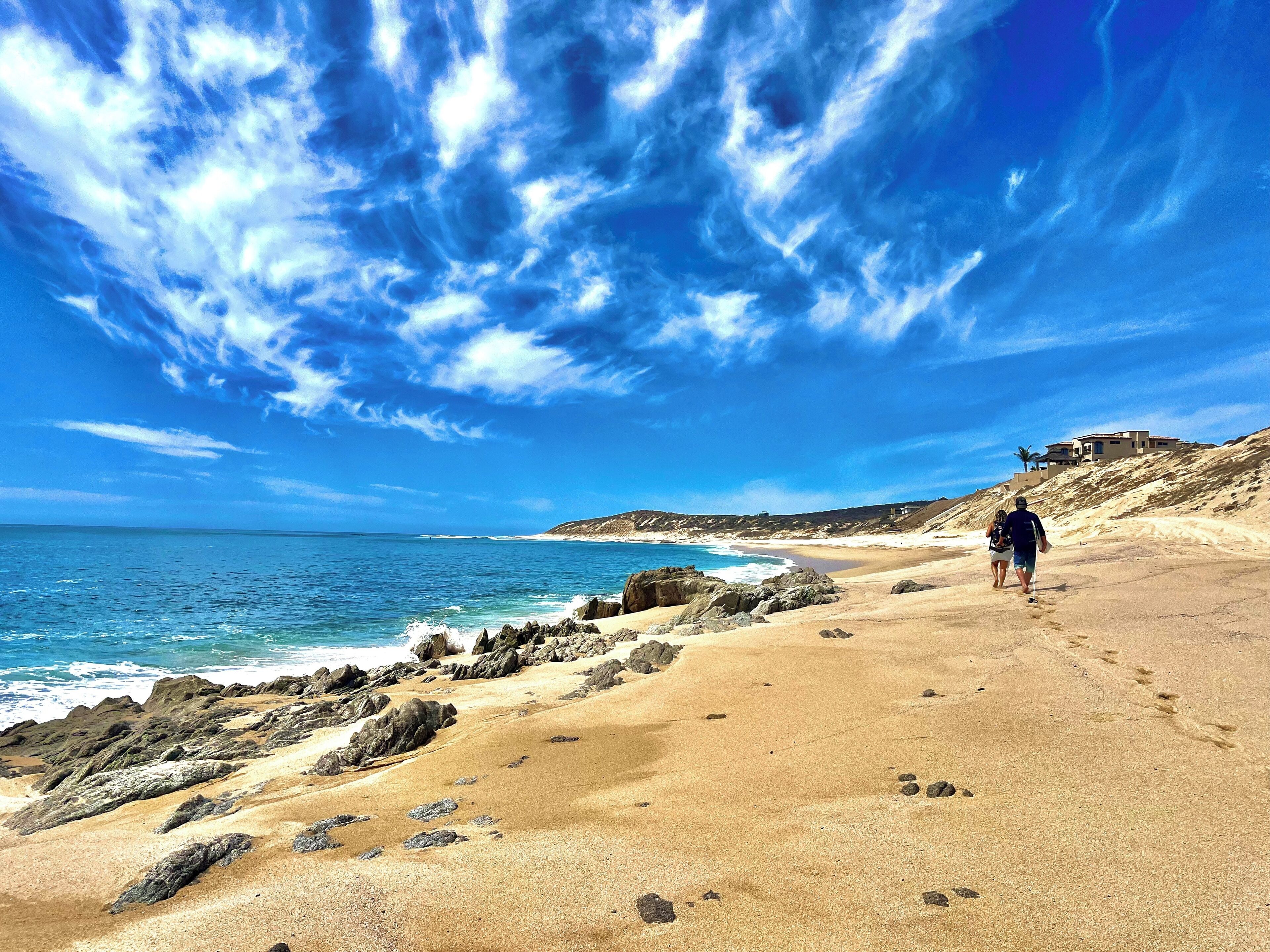 A sandy beach with rocky outcrops, a clear blue sky, and a person walking.
