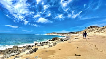 A sandy beach with rocky outcrops, a clear blue sky, and a person walking.