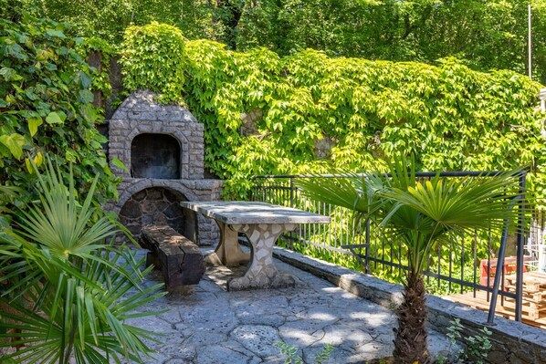 A stone-built outdoor seating area with a stone oven, surrounded by lush greenery and a stone pathway.