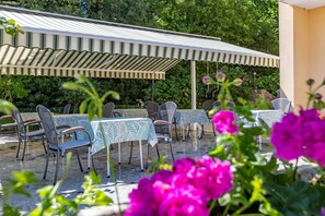 An outdoor patio with tables and chairs, a covered area, and lush greenery in the background.