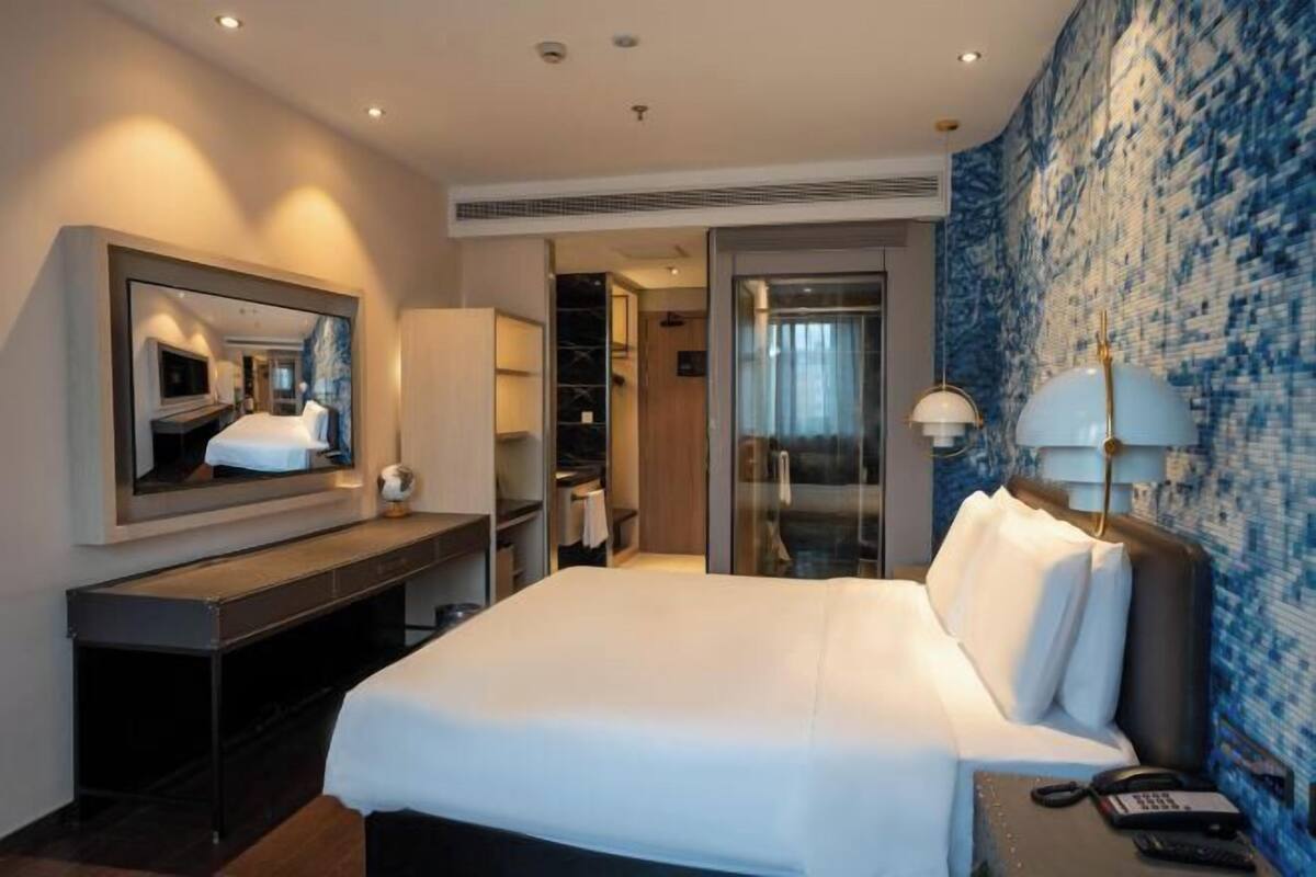 A hotel room with a large bed, a desk with a mirror, a wardrobe, and a bathroom with a glass door.