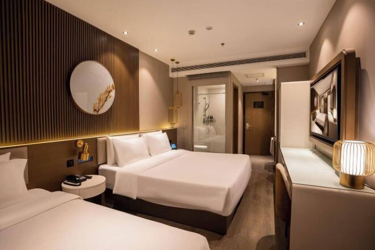 A hotel room with two beds, a round mirror, a bedside table with a lamp, and a view of the bathroom.
