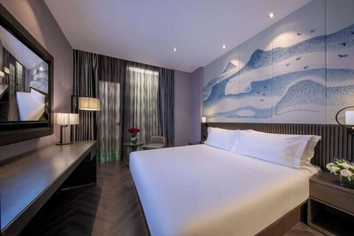 A modern hotel room with a large bed, a desk, a chair, a lamp, and a wall mural.