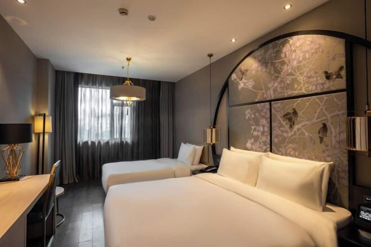 A hotel room with two beds, a desk, a chair, a lamp, and a large window with curtains.