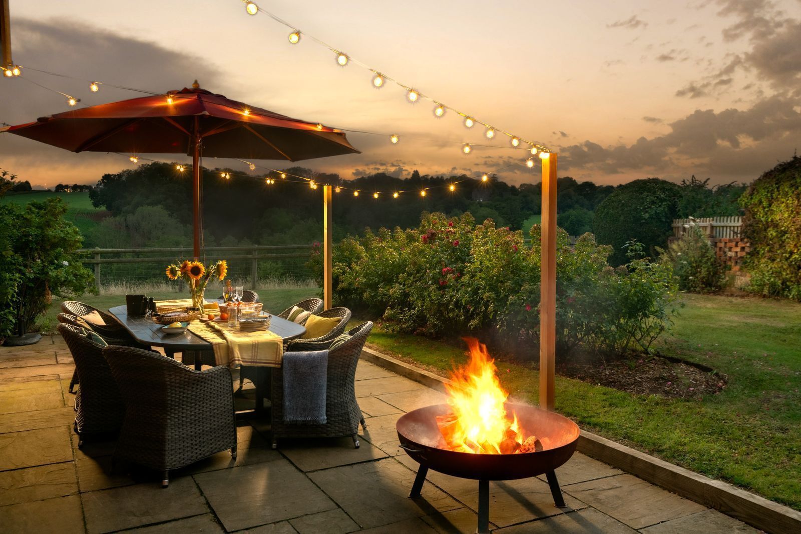An outdoor patio with a dining table, chairs, a fire pit, string lights, and a large umbrella.