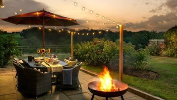 An outdoor patio with a dining table, chairs, a fire pit, string lights, and a large umbrella.
