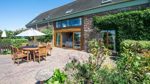 A modern house with a patio area featuring a wooden table and chairs, a large umbrella, and a stone pathway. - Cart Cottage - sleeps 6 guests  in 3 bedrooms (Bridgnorth)