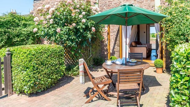Une terrasse avec une table et des chaises en bois, un parasol vert et, au fond, un bâtiment en pierre.