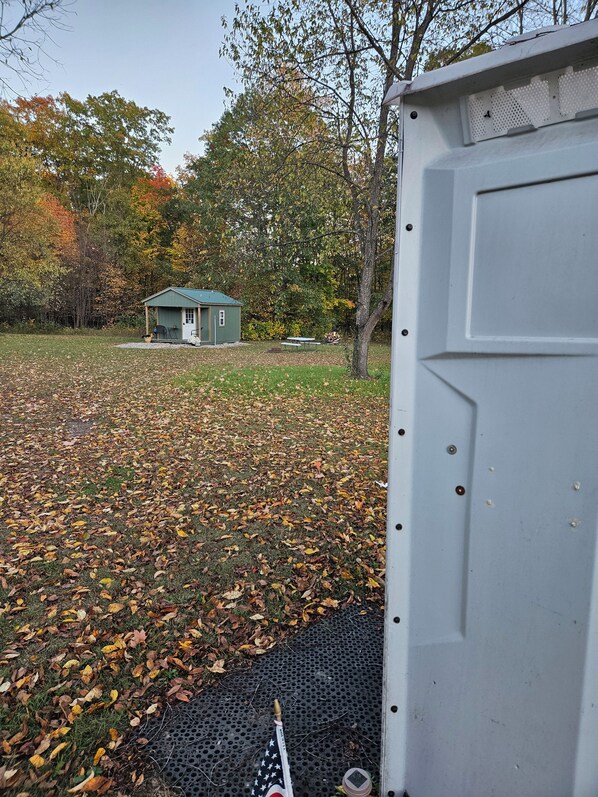 A small cabin with a porch, surrounded by a grassy area with fallen leaves, and a metal structure to the right.