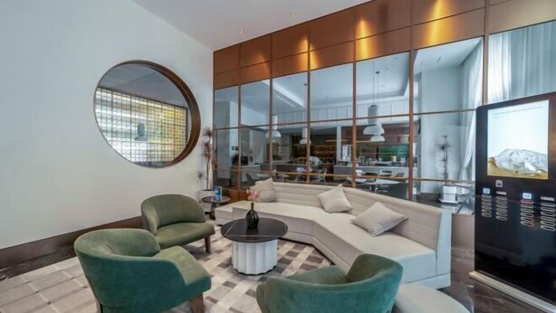 Una sala de estar moderna con un sofá curvo, dos sillones verdes, una mesa redonda de centro y un espejo grande que refleja la habitación.