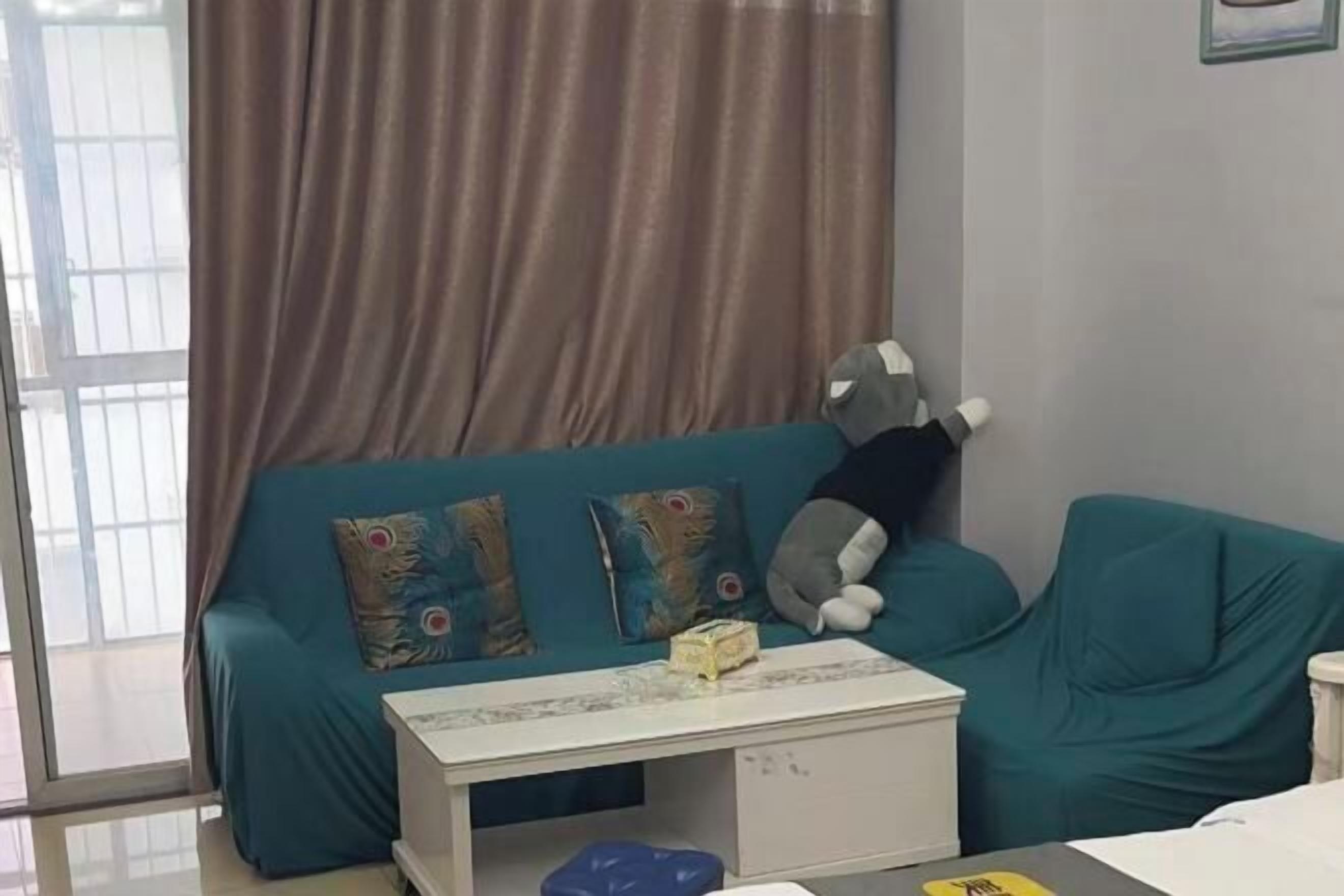 A teal sofa with colorful cushions, a small white table with a box, and a large stuffed animal leaning against the wall.