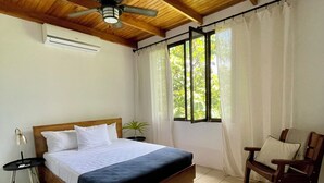Un dormitorio con cama de madera, ventilador de techo, ventana con cortinas, una silla y una pequeña mesa con lámpara.