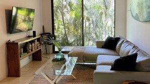 Una sala de estar con televisor, sofá, mesa de centro y un ventanal con vistas a árboles.