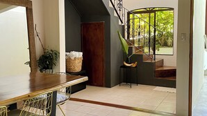Una habitación amplia con techo de madera, zona de comedor con mesa y sillas, una escalera que sube a un nivel superior y vistas a zonas verdes.