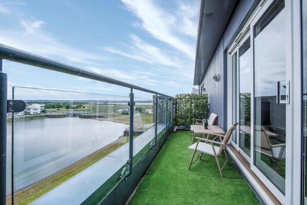 Ein Balkon mit Blick auf einen Fluss und Gebäude, ein kleiner Tisch mit Stühlen und eine Fläche mit grüner Kunstrasen.