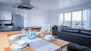 Una cocina moderna con una isla central, un comedor con mesa para cuatro personas y un sofá de cuero negro frente a grandes ventanales.