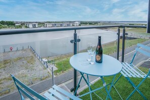 Ein Balkon mit Tisch, zwei Gläsern und einer Weinflasche, mit Blick auf einen Fluss. Im Hintergrund sind Gebäude und eine Brücke zu sehen.
