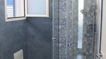 A bathroom with a shower enclosure, a window, and a wooden shelf.